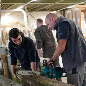 Zwei Handwerker sägen gemeinsam ein Holzbrett in einer modernen Tischlerei-Werkstatt.