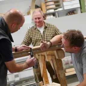 Drei Handwerker messen gemeinsam ein Holzbrett in einer modernen Werkstatt.