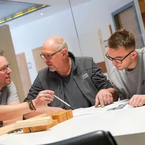 Drei Handwerker besprechen Baupläne und Holzproben in einem modernen Büro.