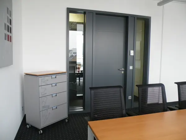Modernes Büro mit Konferenztisch, schwarzen Stühlen und grauem Aktenschrank auf Rollen.